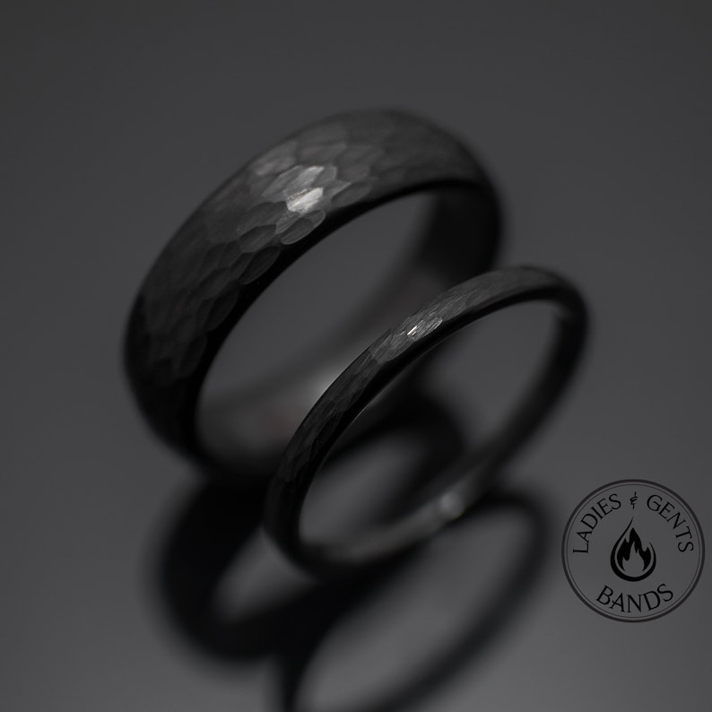 Op de afbeelding: Twee zwarte gehamerde metalen ringen, de ene breder dan de andere, worden getoond tegen een donkere achtergrond. De ringen hebben een getextureerd oppervlak en een cirkelvorm. De afbeelding bevat een logo met de tekst "Ladies & Gents Bands".