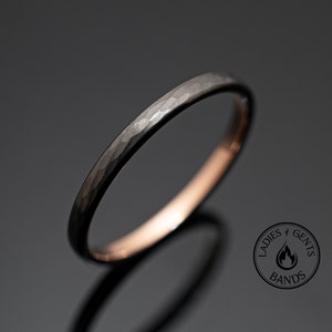 Pode incluir: Um anel preto martelado com uma superfície texturizada. O anel tem uma banda fina e uma cor cinza escuro. O anel está sobre um fundo cinza escuro. O anel é uma joia.