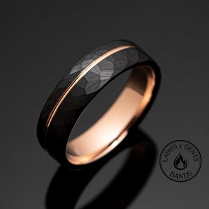 Op de afbeelding: Een zwarte ring met een gehamerde textuur en een dunne, roségouden streep. De binnenkant van de ring is roségoud. De ring staat op een donkere achtergrond. Het is een trouwring.