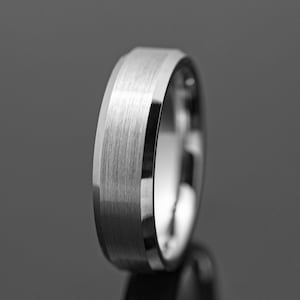 Peut inclure: Gros plan d'une bague argentée avec une bande centrale en métal brossé et des bords polis. La bague a un design moderne et minimaliste, reflétant la lumière et l'ombre. La bague est probablement en acier inoxydable ou en métal similaire.