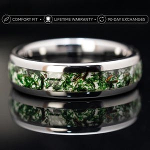 Pode incluir: Um anel de tom prateado polido com uma incrustação central de musgo verde e material branco. Acima do anel, o texto diz: COMFORT FIT, LIFETIME WARRANTY e 90-DAY EXCHANGES. O anel tem uma superfície lisa e reflexiva.