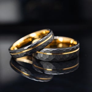 Könnte beinhalten: Zwei schwarze Ringe mit gehammerter Textur und einem dünnen Goldband. Die Innenseite der Ringe ist goldfarben. Die Ringe werden auf einer reflektierenden schwarzen Oberfläche präsentiert.