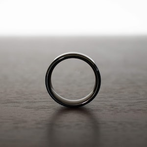 May include: A circular ring with a polished metallic outer surface and a smooth, light-colored inner band. The ring is positioned upright, showcasing its simple, clean profile and reflective finish against a neutral, dark-toned surface.