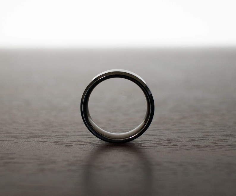 May include: A circular ring with a polished metallic outer surface and a smooth, light-colored inner band. The ring is positioned upright, showcasing its simple, clean profile and reflective finish against a neutral, dark-toned surface.