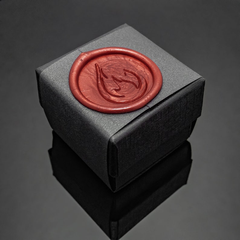 May include: A small, black gift box with a textured surface. A round, red wax seal with a stylized design is affixed to the top. The box is set against a dark, reflective background.