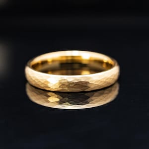 Pode incluir: Um anel de ouro com textura martelada. O anel é redondo e reflete a luz, criando uma aparência brilhante e brilhante. O anel está sobre um fundo escuro.
