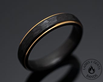 May include: A black ring featuring a textured, hammered finish on the outer surface, accented by a thin, polished gold-tone band running along the center. The ring includes a circular logo with the text LADIES & GENTS BANDS.