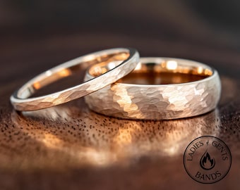 May include: Two rose gold rings with a hammered texture. The thinner ring is positioned at an angle, partially overlapping the wider, thicker ring. Both bands feature a reflective, metallic finish and a circular shape. A circular logo reads LADIES GENTS BANDS.