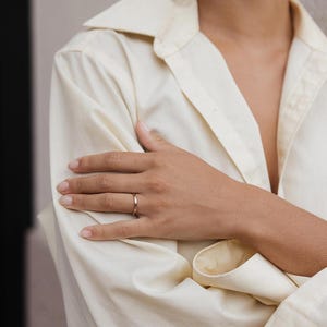 May include: A thin, polished gold band ring with a smooth, rounded surface. The ring features a simple, continuous circular design with a reflective metallic finish, worn on the finger of a hand resting against a light-coloured button-down shirt.