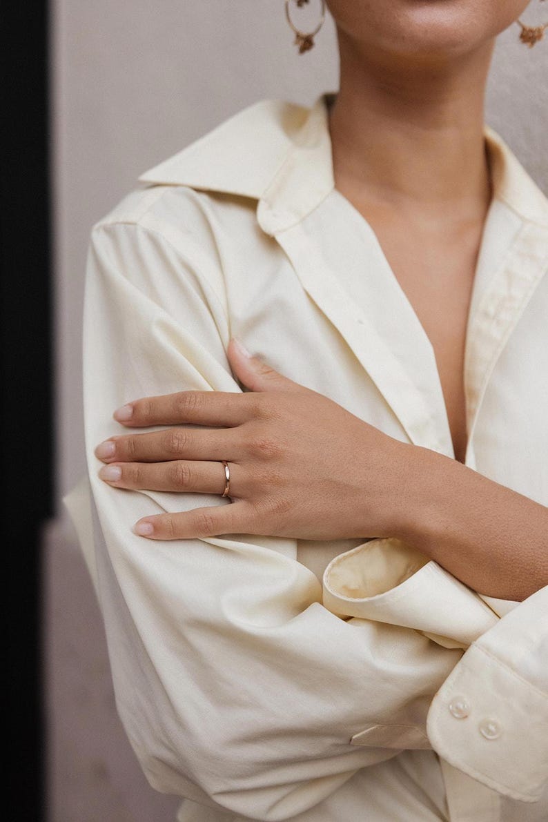 May include: A thin, polished gold band ring with a smooth, rounded surface. The ring features a simple, continuous circular design with a reflective metallic finish, worn on the finger of a hand resting against a light-coloured button-down shirt.