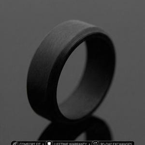 May include: A black ring with a brushed texture, set against a dark background. The ring has a flat top and beveled edges. The image also includes text that reads "COMFORT FIT", "LIFETIME WARRANTY", and "90-DAY EXCHANGES".
