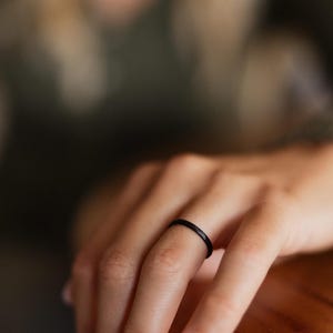 Op de afbeelding: Een dunne, zwarte ring wordt op een vinger gedragen. De ring is een eenvoudige band en de focus ligt op het sieraad. De achtergrond is wazig, met een houten oppervlak en de hand van een persoon zichtbaar.