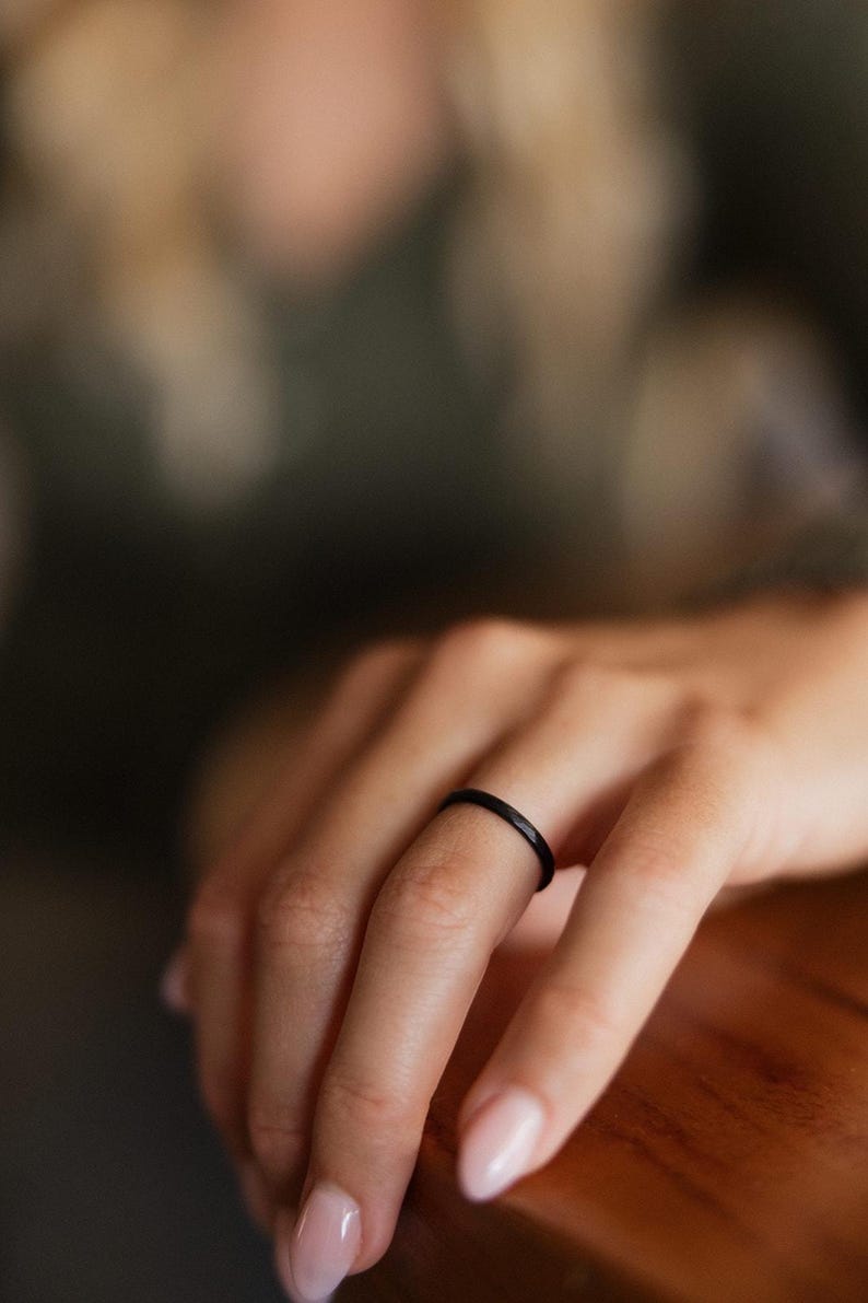 Op de afbeelding: Een dunne, zwarte ring wordt op een vinger gedragen. De ring is een eenvoudige band en de focus ligt op het sieraad. De achtergrond is wazig, met een houten oppervlak en de hand van een persoon zichtbaar.