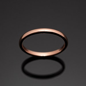May include: A polished rose gold band ring with a smooth, rounded surface. The ring features a simple, continuous circular design with a reflective metallic finish that catches the light, creating a warm, copper-toned appearance against a dark background.