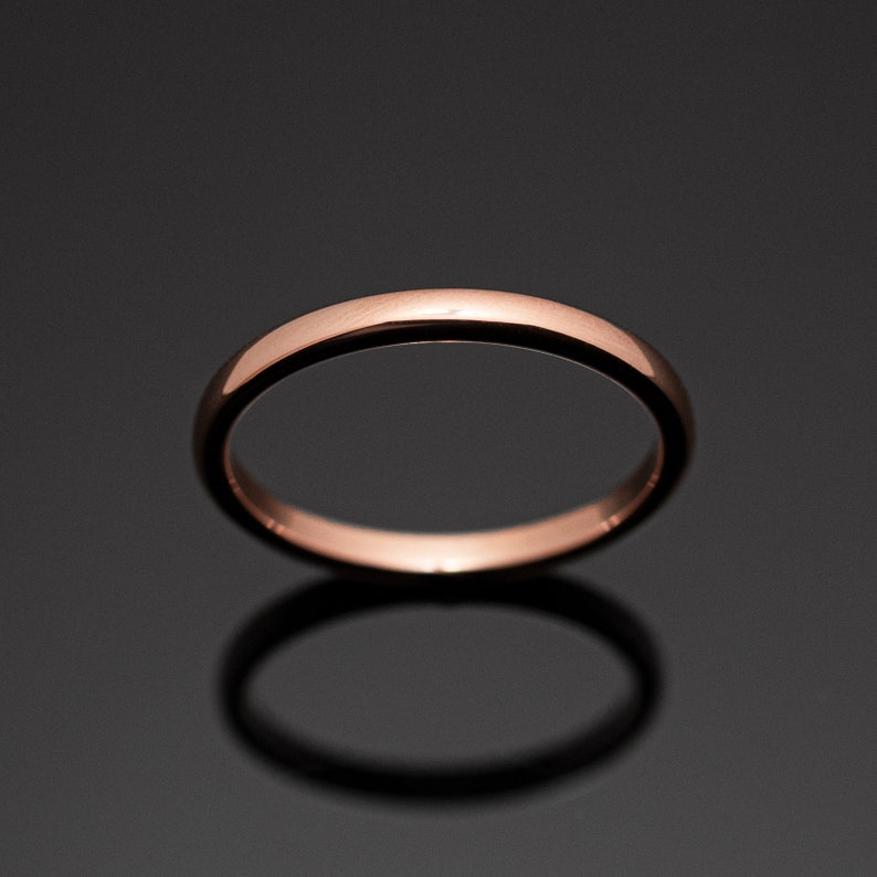 May include: A polished rose gold band ring with a smooth, rounded surface. The ring features a simple, continuous circular design with a reflective metallic finish that catches the light, creating a warm, copper-toned appearance against a dark background.