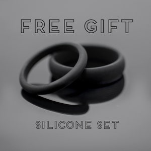 Op de afbeelding: Twee zwarte siliconen ringen, &eacute;&eacute;n dun en &eacute;&eacute;n dikker, liggen op een reflecterend oppervlak. De tekst "FREE GIFT" en "SILICONE SET" staan boven en onder de ringen. De afbeelding heeft een monochrome, minimalistische uitstraling.