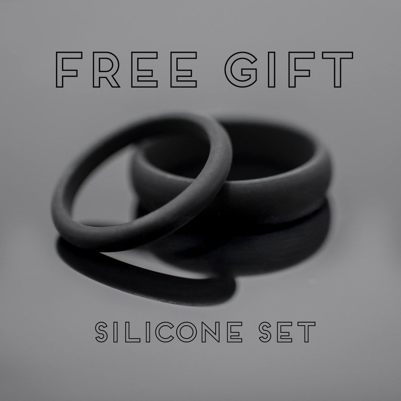 Op de afbeelding: Twee zwarte siliconen ringen, &eacute;&eacute;n dun en &eacute;&eacute;n dikker, liggen op een reflecterend oppervlak. De tekst "FREE GIFT" en "SILICONE SET" staan boven en onder de ringen. De afbeelding heeft een monochrome, minimalistische uitstraling.