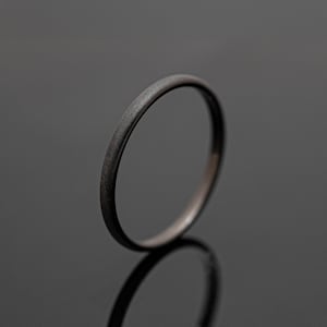 May include: A dark gray ring with a matte finish. The ring is a simple band design, with a smooth, rounded surface. The ring is displayed on a reflective surface, creating a mirrored effect. The ring appears to be made of metal.