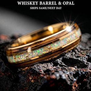 May include: A gold ring with a whiskey barrel and opal design. The ring features a band of dark wood, a center of iridescent opal, and gold accents. The text "WHISKEY BARREL & OPAL" is at the top.