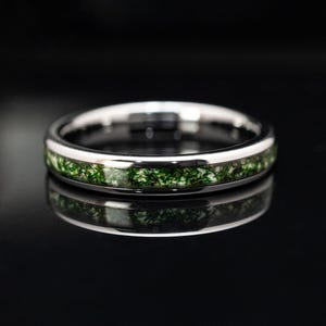 May include: A silver ring with a band of green moss-like material. The ring is set against a black background, with a reflection of the ring visible below. The ring is a close-up shot, highlighting the details of the design.