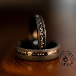 Könnte beinhalten: Zwei schwarze Eheringe mit strukturiertem, gesprenkeltem Inlay. Die Ringe liegen auf einer Holzoberfläche, wobei ein Ring im Fokus steht und der andere leicht verschwommen im Hintergrund. Die Ringe haben ein mattes Finish.