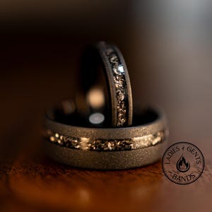 May include: Two dark gray wedding bands with a textured finish. Each ring features a band of sparkling, light-colored material. The rings are stacked, with one slightly behind the other. The logo for "Ladies & Gents Bands" is visible.