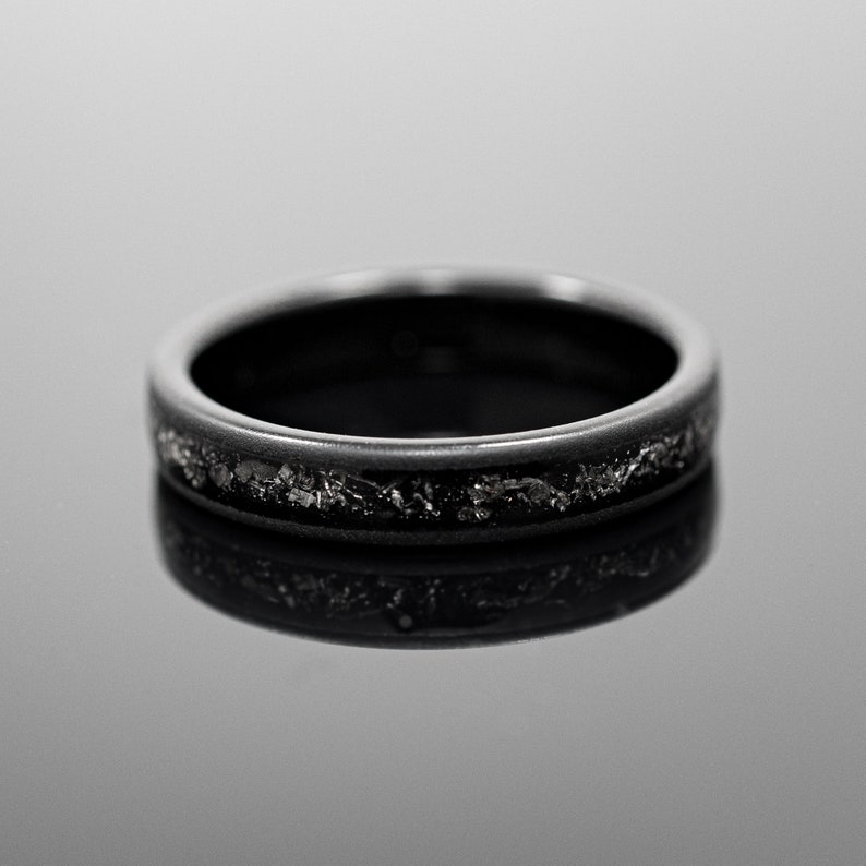 Peut inclure: Une bague noire avec une bande de mat&eacute;riau gris fonc&eacute; textur&eacute;. La bague est pos&eacute;e sur une surface r&eacute;fl&eacute;chissante, soulignant sa finition polie. Le design de la bague est simple, mais &eacute;l&eacute;gant, adapt&eacute; au port quotidien.