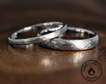 May include: Two silver-toned rings with a hammered, textured finish. The rings are positioned one behind the other. A circular logo in the bottom right corner features a flame icon and the text LADIES GENTS BANDS.