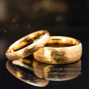Two gold wedding rings with a textured finish are displayed on a reflective surface against a dark background.