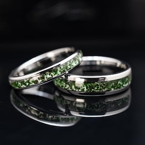 May include: Two silver-toned metal rings featuring a central inlay of green moss-like material. The rings have a polished, reflective finish and are positioned at an angle, showing the detailed texture of the green organic inlay.