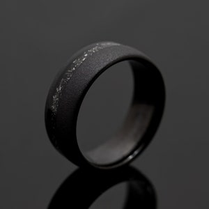 May include: A black ring with a textured surface and a silver inlay. The ring is made of a dark material and has a simple, elegant design. The ring is shown on a reflective surface.