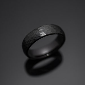 Op de afbeelding: Een zwarte ring met een getextureerde, gehamerde afwerking. De ring heeft een afgeronde band en een donker, reflecterend oppervlak. De ring is een sieraad, waarschijnlijk voor een bruiloft of verloving.