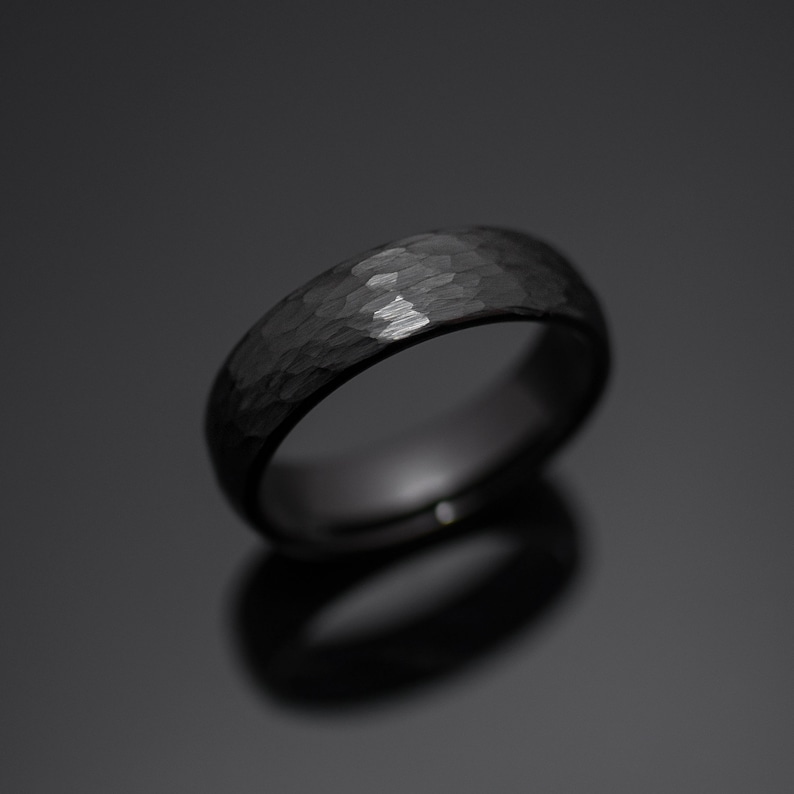 Op de afbeelding: Een zwarte ring met een getextureerde, gehamerde afwerking. De ring heeft een afgeronde band en een donker, reflecterend oppervlak. De ring is een sieraad, waarschijnlijk voor een bruiloft of verloving.