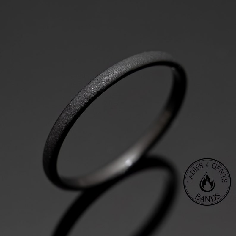 May include: A close-up shot of a black ring with a textured surface. The ring is thin and has a matte finish. The ring is set against a dark background. The ring is a band, and the image includes the text "Ladies & Gents Bands".