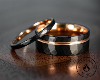 May include: Two wedding bands. The larger ring is black with a hammered texture and a rose gold interior and center band. The smaller ring is black with a hammered texture and a rose gold interior. The rings are on a wooden surface.