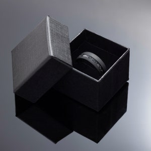 May include: A black ring with a silver inlay is displayed inside an open black gift box. The ring has a dark matte finish and a band of silver-colored material. The box is rectangular with a textured surface.
