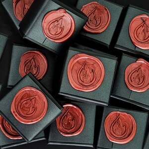 May include: An overhead shot shows several black gift boxes, each sealed with a reddish-brown wax seal. The seals feature a stylized flame design. The boxes are arranged closely together against a dark background, creating a pattern.
