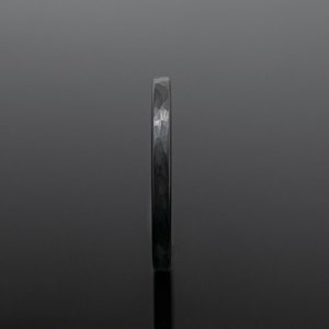 Op de afbeelding: Een donkergrijze gehamerde metalen ring. De ring heeft een textuuroppervlak en een smalle band. De ring wordt gepresenteerd tegen een donkere achtergrond, wat de metalen glans benadrukt. Dit sieraad is geschikt voor dagelijks gebruik.
