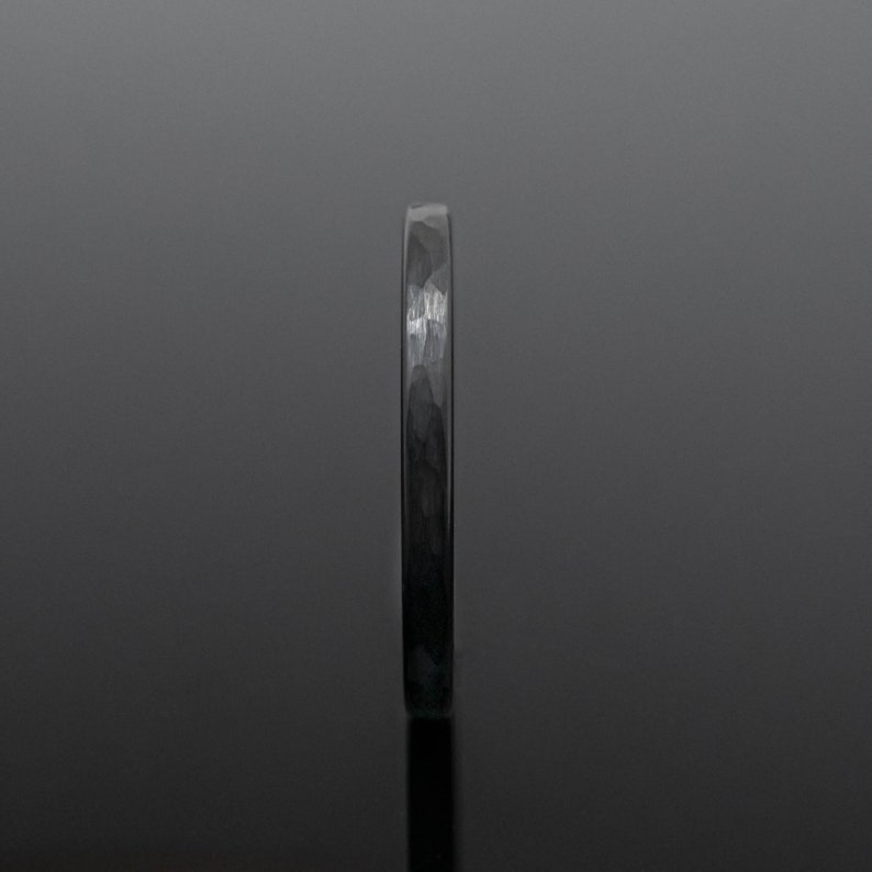 Op de afbeelding: Een donkergrijze gehamerde metalen ring. De ring heeft een textuuroppervlak en een smalle band. De ring wordt gepresenteerd tegen een donkere achtergrond, wat de metalen glans benadrukt. Dit sieraad is geschikt voor dagelijks gebruik.