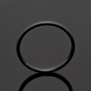 May include: A matte black ring with a textured surface. The ring is circular and reflects on the dark surface below. The ring is a simple band, suitable for everyday wear.