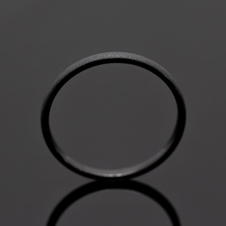 May include: A matte black ring with a textured surface. The ring is circular and reflects on the dark surface below. The ring is a simple band, suitable for everyday wear.