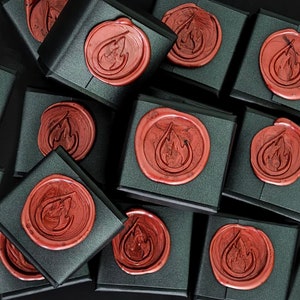 May include: A collection of square black gift boxes, each sealed with a circular red wax seal featuring an embossed flame icon. The boxes are arranged in a cluster, showcasing the consistent matte black finish and the glossy red wax.