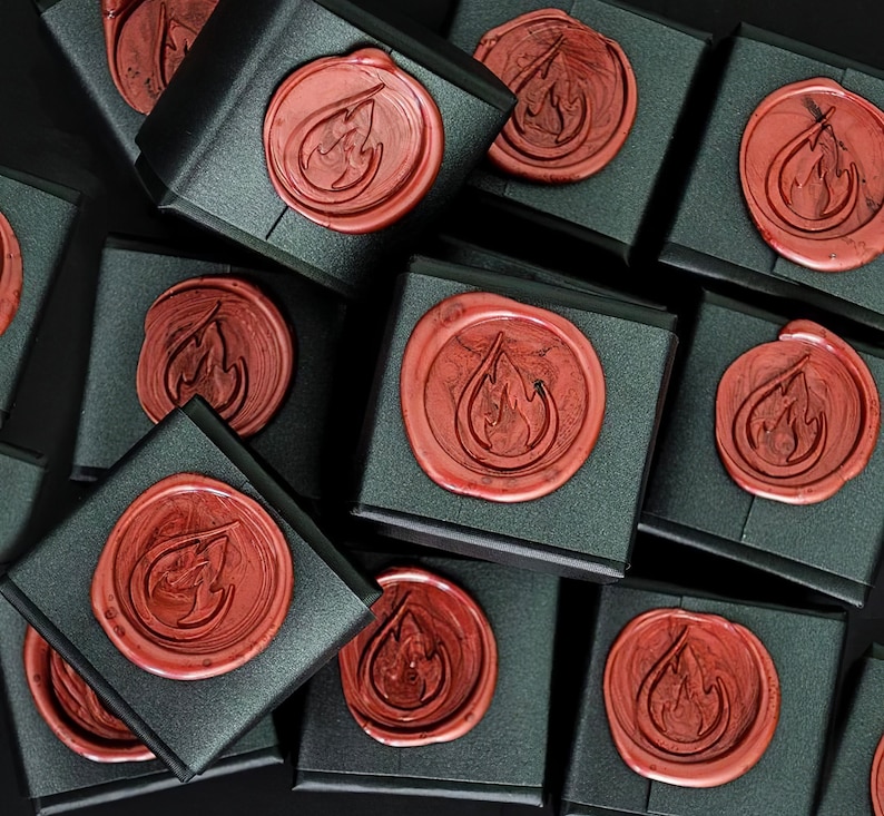 May include: A collection of square black gift boxes, each sealed with a circular red wax seal featuring an embossed flame icon. The boxes are arranged in a cluster, showcasing the consistent matte black finish and the glossy red wax.