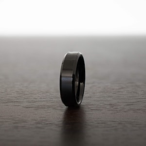 May include: A solid black ring with a smooth, polished finish and beveled edges. The band has a uniform width and a reflective surface that catches the light, showcasing a sleek, dark metallic appearance in a simple, circular design.