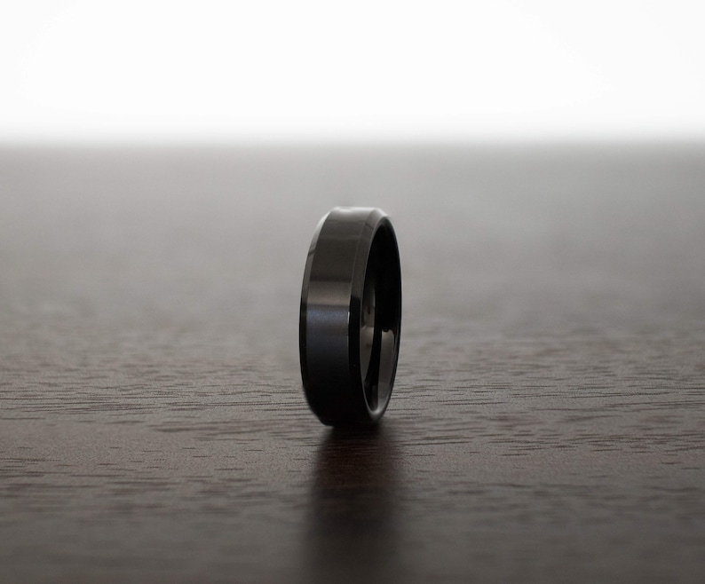 May include: A solid black ring with a smooth, polished finish and beveled edges. The band has a uniform width and a reflective surface that catches the light, showcasing a sleek, dark metallic appearance in a simple, circular design.