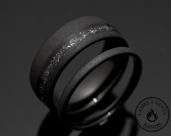 May include: Two black rings, one with a textured surface and a silver inlay. The other ring is smooth and encircles the first. The rings are set against a dark background, with a logo in the lower right corner.
