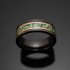 May include: A black band ring featuring a central inlay of green moss-like material bordered by two thin rose gold-colored metallic stripes. The ring has a smooth, polished finish and a rounded profile, showcasing a dark exterior and interior.