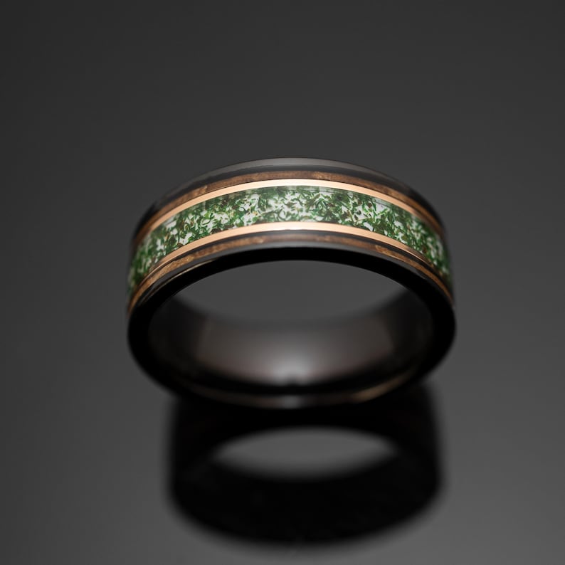 May include: A black band ring featuring a central inlay of green moss-like material bordered by two thin rose gold-colored metallic stripes. The ring has a smooth, polished finish and a rounded profile, showcasing a dark exterior and interior.