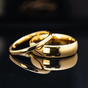 May include: Two gold wedding rings, one thin and one thicker, are displayed on a reflective black surface. The rings are highly polished, reflecting light and creating a bright, elegant appearance. The image highlights the smooth, rounded design of the bands.