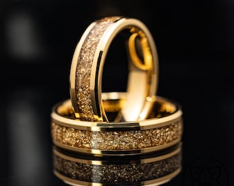Puede incluir: Un anillo de tono dorado con una incrustación central de escamas texturizadas y brillantes de color oro. La banda tiene bordes pulidos y lisos que enmarcan el centro metálico granulado. El diseño es consistente y circular.